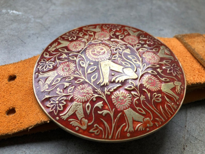 May include: A round, red metal belt buckle with a gold design of stylised flowers and figures. The buckle is attached to a brown leather belt.