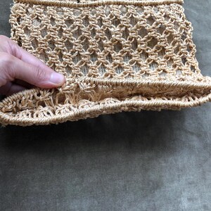 May include: A close-up of a beige, woven bag with a textured, open-weave design. The bag's structure is visible, showcasing the intricate knotting pattern. The bottom edge has a ruffled detail. The bag is held by a hand.