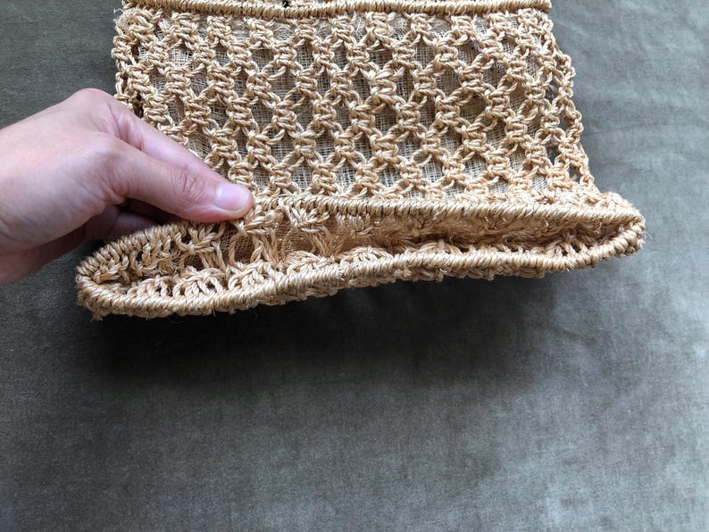 May include: A close-up of a beige, woven bag with a textured, open-weave design. The bag's structure is visible, showcasing the intricate knotting pattern. The bottom edge has a ruffled detail. The bag is held by a hand.