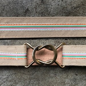 May include: A beige canvas belt with a gold metal buckle. The buckle is a twisted circle design. The belt has a multi-colored stripe pattern in orange, green, white, and pink.