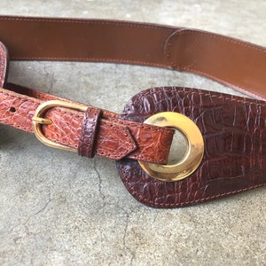 May include: A brown leather belt with a gold buckle and a large, round, gold grommet. The belt has a crocodile-embossed pattern.