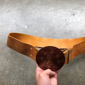 May include: A brown leather belt with a large, round, metal buckle. The buckle features a detailed design of flowers and leaves in red and gold.