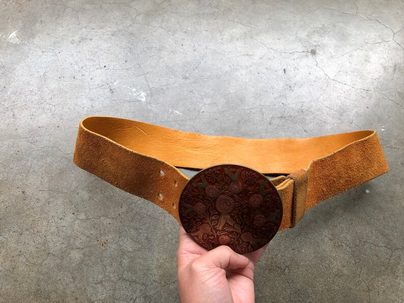 May include: A brown leather belt with a large, round, metal buckle. The buckle features a detailed design of flowers and leaves in red and gold.