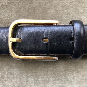May include: Close-up of a black leather belt with a gold-colored rectangular buckle. The belt has several holes for adjustment and a loop to secure the excess length. The buckle is slightly worn, showing signs of use.