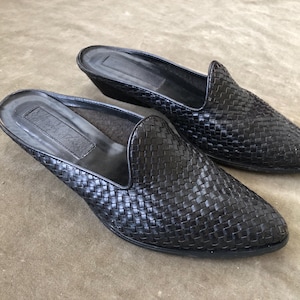 Könnte beinhalten: Ein Paar schwarze, gewebte Lederschlupfschuhe. Die Schuhe haben einen kleinen Absatz und eine runde Spitze. Das Obermaterial besteht aus einem Korbgeflechtmuster. Die Schuhe sind so konzipiert, dass man sie leicht an- und ausziehen kann.