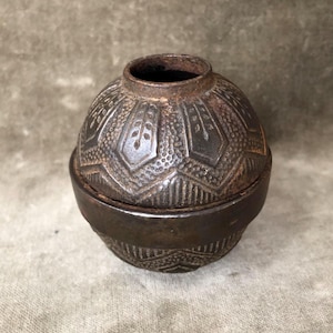 May include: A small, spherical, dark brown metal container with a textured surface featuring repeating geometric patterns and leaf-like motifs. The object consists of two interlocking parts, with a circular opening at the top and a smooth band.