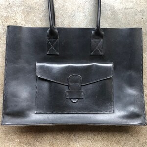 Puede incluir: Un bolso tote de cuero negro con un bolsillo frontal grande y dos asas superiores.