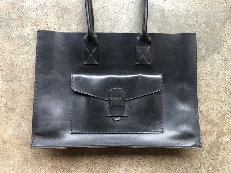 Puede incluir: Un bolso tote de cuero negro con un bolsillo frontal grande y dos asas superiores.