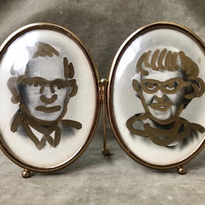 Könnte beinhalten: Ein goldfarbener, doppelt ovaler Bilderrahmen. Jedes Oval enthält ein Schwarz-Weiß-Porträt mit goldfarbener Linienkunst. Die Porträts zeigen einen Mann und eine Frau, beide mit Brille. Der Rahmen ist klappbar.