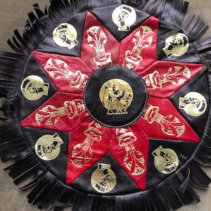 May include: A round, black and red leather decorative piece with gold accents. The design features a central circle with a camel illustration, surrounded by a starburst pattern and gold medallions. The outer edge has a fringe.