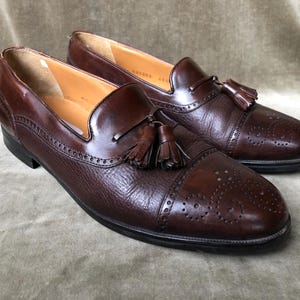 May include: A pair of brown leather tassel loafers with a brogue pattern. The loafers have a pointed toe and a tassel detail on the vamp.