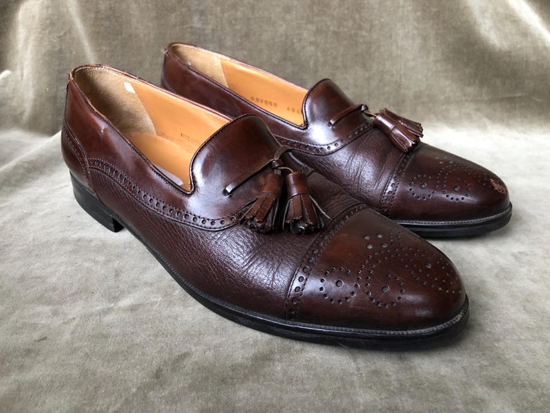 May include: A pair of brown leather tassel loafers with a brogue pattern. The loafers have a pointed toe and a tassel detail on the vamp.