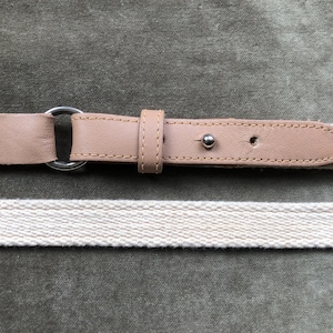 Puede incluir: Un cinturón de cuero marrón con una hebilla plateada y una correa de algodón blanco. El cinturón está sobre un fondo de tela verde.