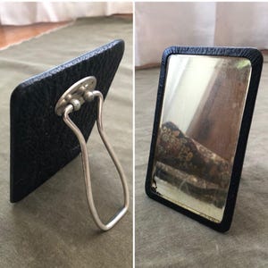 Puede incluir: Un pequeño espejo de tocador rectangular con marco y respaldo de símil piel negro. El espejo tiene un soporte articulado plateado. El espejo refleja una imagen borrosa del entorno. Es un artículo vintage.