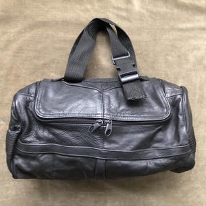 Puede incluir: Bolso de mano de cuero negro con asa superior y bandolera. El bolso presenta un cierre de solapa, dos cremalleras y un detalle de hebilla. El cuero tiene un aspecto texturizado.