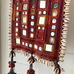 Può includere: Un arazzo rettangolare con una base in tessuto rosso scuro e arancione decorata con numerosi piccoli specchi quadrati. I bordi sono rifiniti con conchiglie di ciprea bianche e il fondo presenta tre nappe decorative con conchiglie.