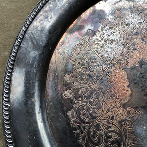May include: Antique silver-plated serving tray with ornate floral and scrollwork design. The tray has a detailed, raised edge and a tarnished finish, showing signs of age and use. The intricate pattern covers a large portion of the tray's surface.