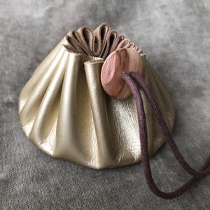 May include: A gold leather pouch with a drawstring. It features a wooden button and a brown drawstring. The pouch is gathered at the top, giving it a soft, textured look. The background is a neutral grey.