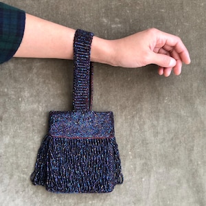 May include: A small, square-shaped handbag covered in dark, iridescent beads that shimmer with blue, purple, and red tones. The bag features a single, matching beaded wrist strap and a dense layer of long, dangling beaded fringe at the base.