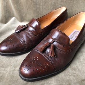 May include: A pair of brown leather tassel loafers with a wingtip design and a perforated pattern. The loafers have a brown leather tassel on each shoe.