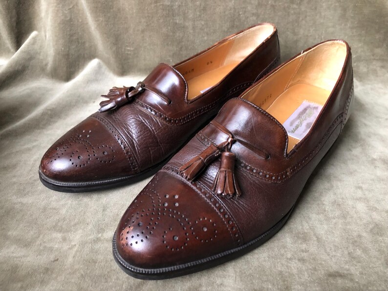 May include: A pair of brown leather tassel loafers with a wingtip design and a perforated pattern. The loafers have a brown leather tassel on each shoe.
