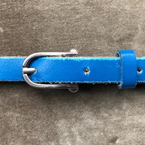May include: A close-up of a bright blue leather belt with a silver-colored buckle. The belt has a thin profile and is secured with a classic buckle. The belt is approximately 0.5 inches wide.