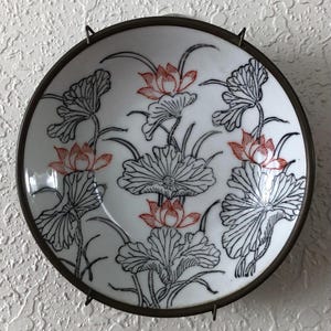 May include: A white ceramic plate with a floral design featuring red lotus flowers and black line art leaves. The plate has a dark metal rim and is mounted on a wall. The design is reminiscent of Asian art.