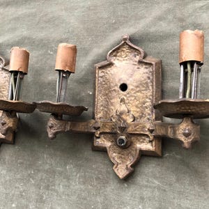 May include: Three antique brass wall sconces with ornate detailing. Each sconce features a decorative backplate, a small dish, and a socket with a brown paper sleeve. The sconces are mounted on a green surface.