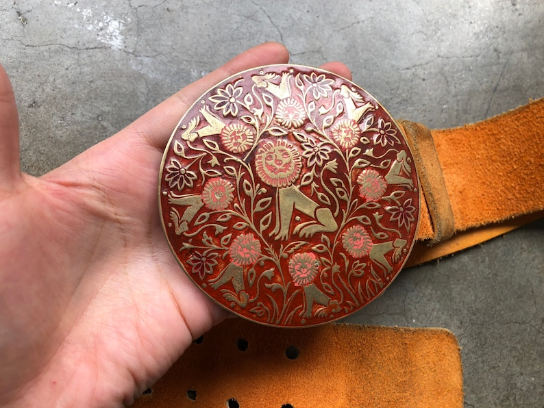 May include: A round, red and gold belt buckle with a detailed design of stylised lions and flowers. The buckle is attached to a brown leather belt.
