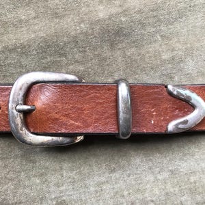 May include: A brown leather belt with a silver buckle and a silver tip. The buckle is a simple oval shape, and the tip is a pointed oval shape.