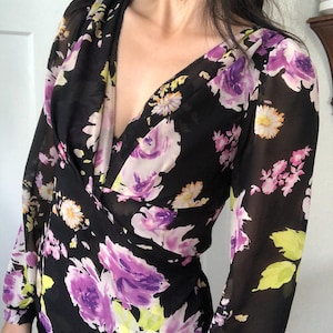 Peut inclure: Haut noir de style cache-cœur à manches longues, orné d'un motif floral. Le design présente de grandes fleurs violettes et roses, avec des accents jaunes et blancs, sur un tissu noir transparent.