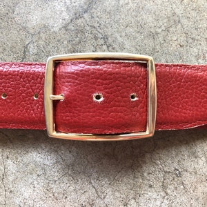 May include: A red leather belt with a gold metal buckle. The belt is made of a textured leather and has a simple design.