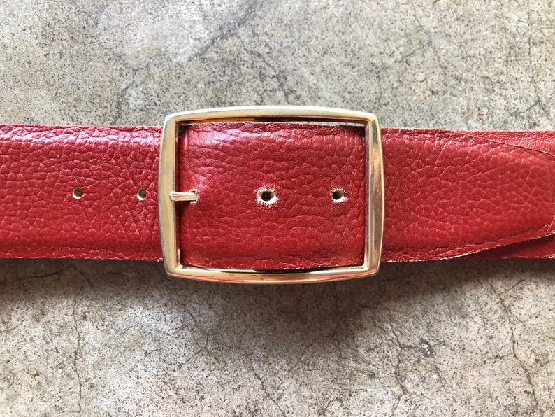 May include: A red leather belt with a gold metal buckle. The belt is made of a textured leather and has a simple design.