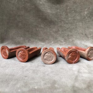 May include: Five terracotta-colored ceramic objects, each featuring a cylindrical base with a circular face embossed with a detailed floral design and a long, narrow, grooved body extending horizontally from the circular end.