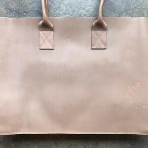 May include: A light brown leather tote bag with two handles. The bag has a subtle, worn look and is made of a soft, supple leather.