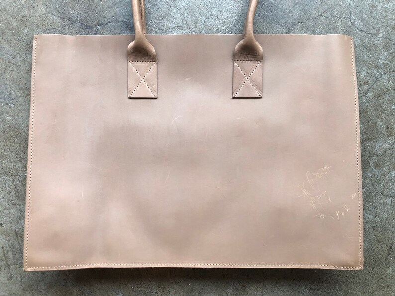 May include: A light brown leather tote bag with two handles. The bag has a subtle, worn look and is made of a soft, supple leather.
