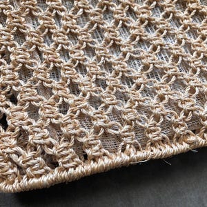 May include: Close-up of a beige macrame textile with a diamond pattern. The woven design is made of thick, natural-colored fibers, creating a textured surface. The edges are neatly finished, showcasing the craftsmanship of the piece.