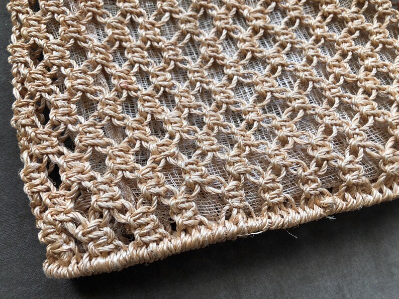 May include: Close-up of a beige macrame textile with a diamond pattern. The woven design is made of thick, natural-colored fibers, creating a textured surface. The edges are neatly finished, showcasing the craftsmanship of the piece.