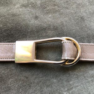 Puede incluir: Un cinturón de cuero marrón claro con una hebilla dorada. La hebilla tiene un diseño único con un doble bucle.