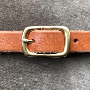 May include: Close-up of a tan leather belt with a gold-colored rectangular buckle. The belt has two holes and a rough, unfinished edge. The buckle is centered on the belt, and the leather appears to be new.