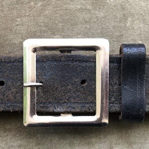 May include: Close-up of a black leather belt with a silver square buckle. The belt has two holes and a loop to secure the belt end. The leather has a worn, textured appearance.
