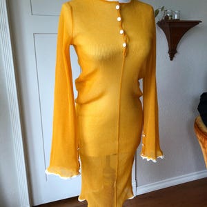 May include: A long-sleeved, yellow dress with a button-up front and a ribbed texture. The dress has a slight flare at the hem and cuffs, trimmed with white scalloped edges. The dress is displayed on a mannequin.