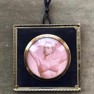 May include: A square wall-hanging frame with a dark blue leather-like backing and a gold-toned metal border. A circular gold frame sits in the center, enclosing a sepia-toned photograph of a person wearing a head wrap and a bandeau top.