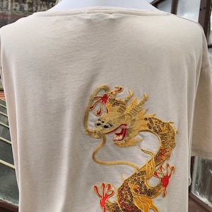 May include: Beige t-shirt featuring an embroidered golden dragon design on the back. The dragon has red accents and intricate detailing. The shirt has a wide neckline and short sleeves, made of a soft, neutral-toned fabric.