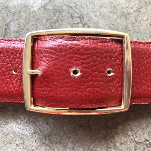 May include: A red leather belt with a gold metal buckle. The buckle is rectangular with rounded corners.
