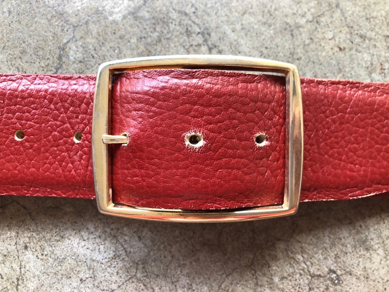 May include: A red leather belt with a gold metal buckle. The buckle is rectangular with rounded corners.