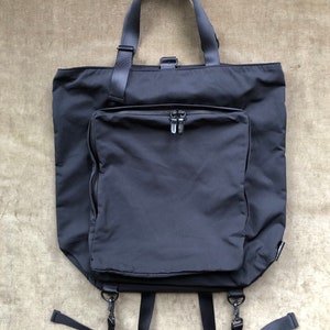 Op de afbeelding: Een zwarte tote bag met een groot ritsvak aan de voorkant. De tas heeft verstelbare banden die als rugzak of schoudertas kunnen worden gedragen.