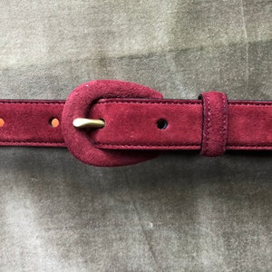 May include: A burgundy suede belt with a large oval buckle. The buckle is made of suede and has a gold metal clasp.