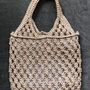 May include: A beige macrame tote bag with a woven, openwork design. The bag features a rectangular body and a sturdy handle. The natural-toned bag is made of a woven material, perfect for carrying items. The bag's dimensions are not visible.