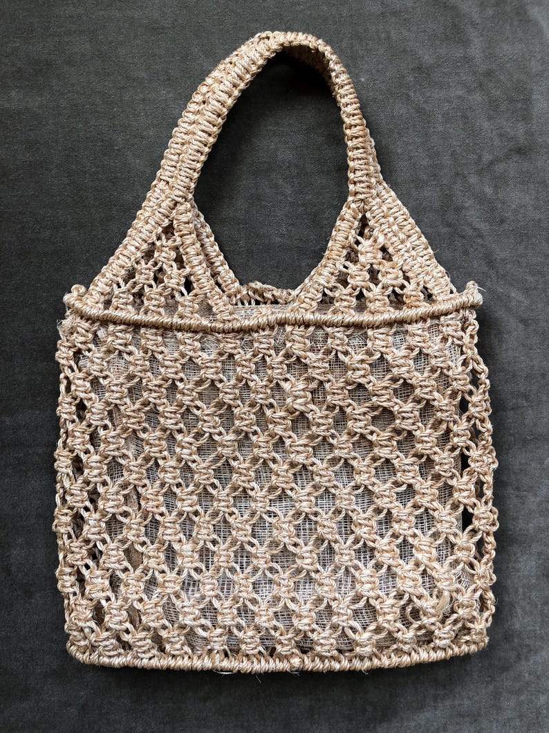 May include: A beige macrame tote bag with a woven, openwork design. The bag features a rectangular body and a sturdy handle. The natural-toned bag is made of a woven material, perfect for carrying items. The bag's dimensions are not visible.